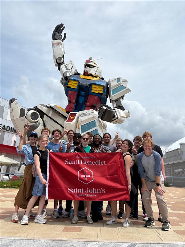 A group of smiling people posing in front of a giant Gundam robot statue, holding a red Saint Benedict and Saint John's University banner on a cloudy day.