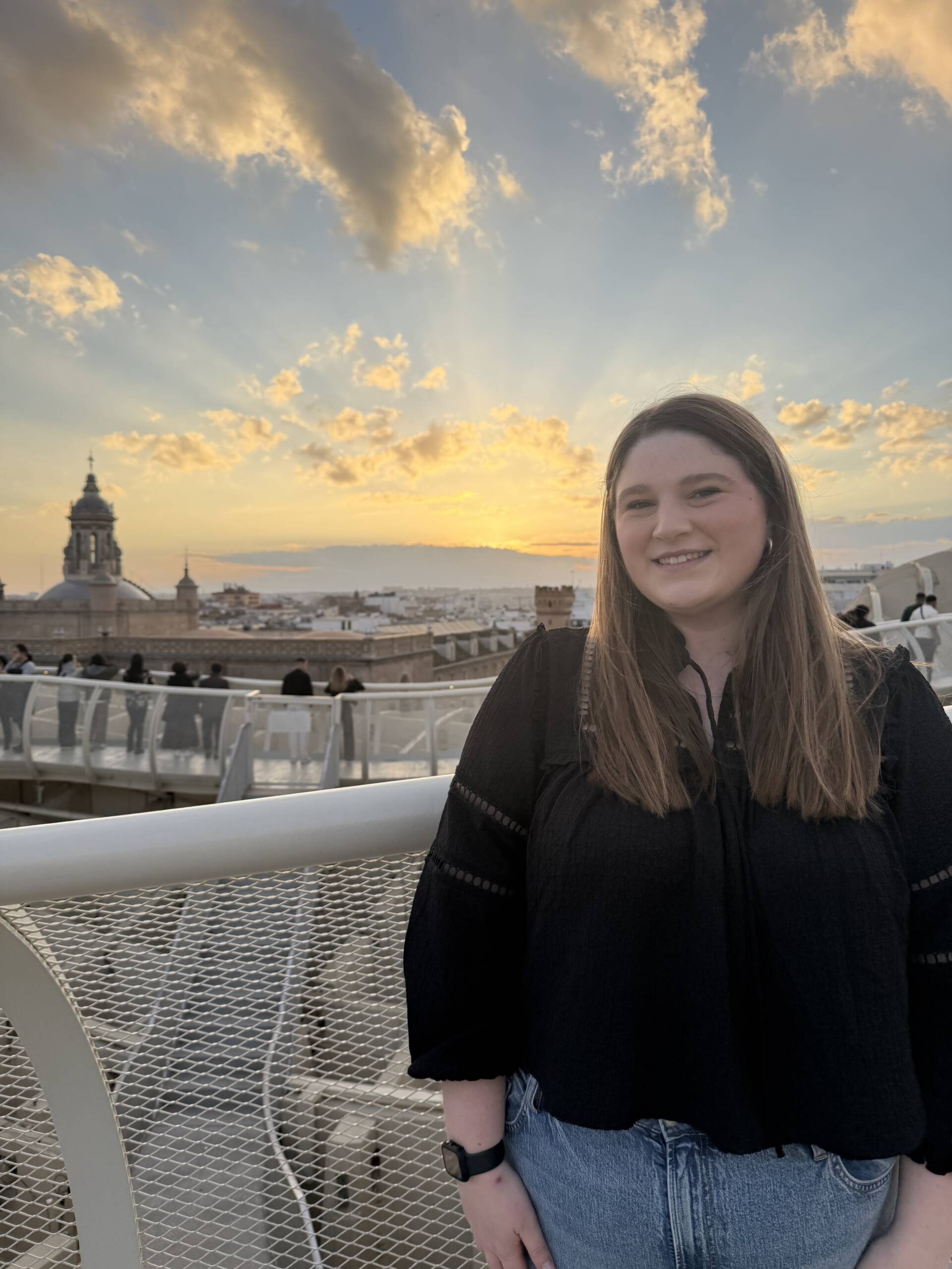 A young woman with long brown hair, wearing a black top and blue jeans, smiles while standing on a balcony at sunset. The cityscape and a church tower are visible in the background under a partly cloudy sky.
