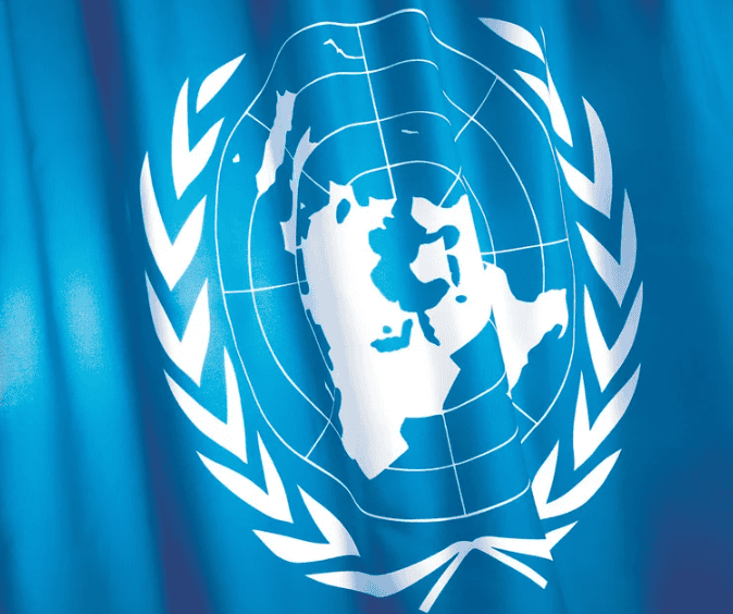 A close-up of the United Nations flag, featuring a white world map centered on the North Pole, surrounded by two olive branches, set against a blue background. The fabric appears to be gently rippling.