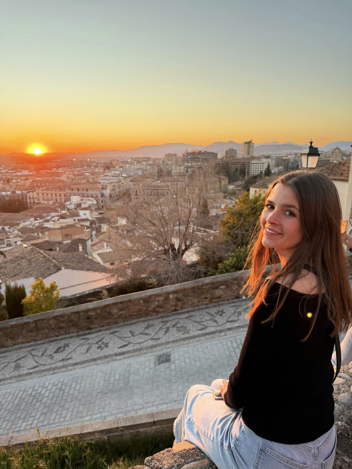 A young woman with long brown hair, wearing a black top and light blue jeans, sits on a stone ledge overlooking a city at sunset, with buildings, mountains, and an orange sky in the background.