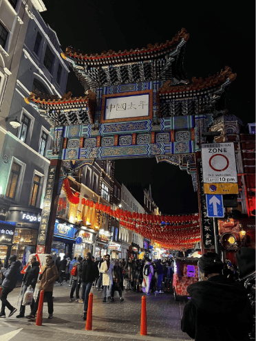 A bustling street scene at night in Chinatown, with a large ornate Chinese gate, red lanterns hanging above, and crowds of people walking beneath brightly lit shopfronts.