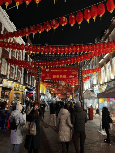 A lively street at night is decorated with red lanterns and a festive banner celebrating Chinese New Year. People in winter clothing walk and take photos under the lights and lanterns.