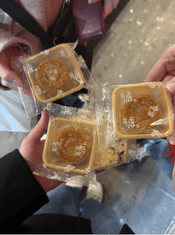 Four hands are holding individually wrapped mooncakes in clear plastic packaging. The people are standing on a tiled floor, and their clothing is partially visible in the background.