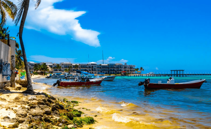 Small boats are anchored near a sandy, palm-lined shore with rocky edges. In the background, there are buildings, a pier, and a clear blue sky with a few clouds. The sea is calm and vibrant under bright sunlight.