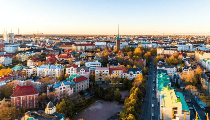 Aerial view of a cityscape with colorful buildings, tree-lined streets, a church with a tall spire in the center, and a clear sky at sunset.