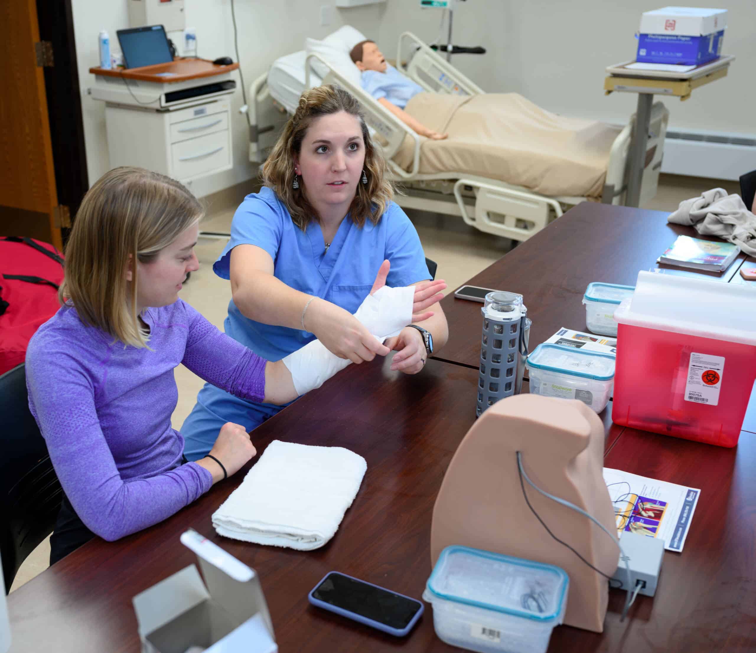 Two women in a medical training room; one is sitting, having her forearm wrapped in a bandage by the other, who is wearing blue scrubs. A medical mannequin lies in a hospital bed in the background. Various medical supplies are scattered on the table in the foreground.