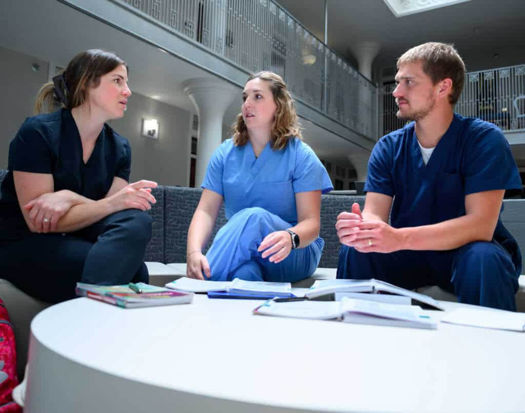 Three people in scrubs sit around a white table with open books and notebooks, engaged in conversation in a bright, modern indoor space with railings and columns in the background.