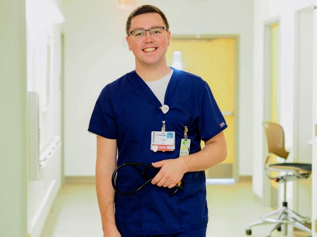 A person wearing blue medical scrubs and an ID badge stands smiling in a brightly lit hallway, holding a stethoscope in one hand.