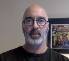A bald man with a white beard wearing glasses is looking straight at the camera. He is indoors with a framed picture on the wall behind him.