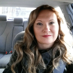 A person with long wavy hair is seated in the driver's seat of a car, wearing a dark jacket and a seatbelt. The background shows the car's interior and a hint of the outside through the windows.