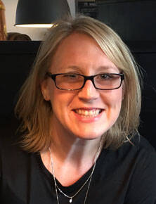 A person with shoulder-length blonde hair and glasses is smiling at the camera. They are wearing a black shirt and a silver necklace. The background includes a black surface and part of a lamp.