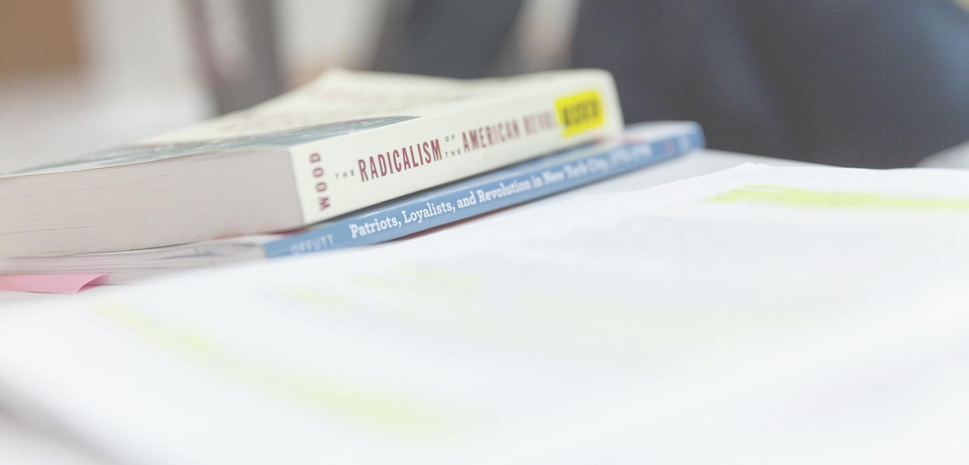 A close-up of a stack of books on a desk. The top book is titled "THE RADICALISM of the AMERICAN REVOLUTION" by Wood, and the second book appears to be about Patriots, Loyalists, and Revolution in New York City. Some papers are scattered in the foreground.