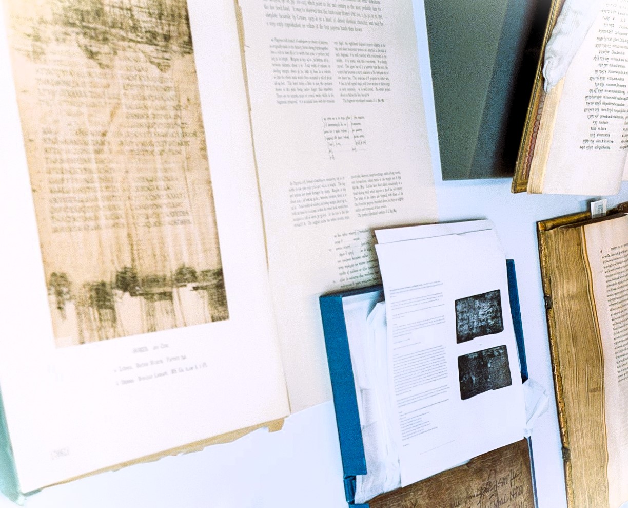 A display of historical documents and pages, some in a folder and others mounted on a wall. The texts appear aged, with printed and handwritten elements, including visible images and tables on the pages.
