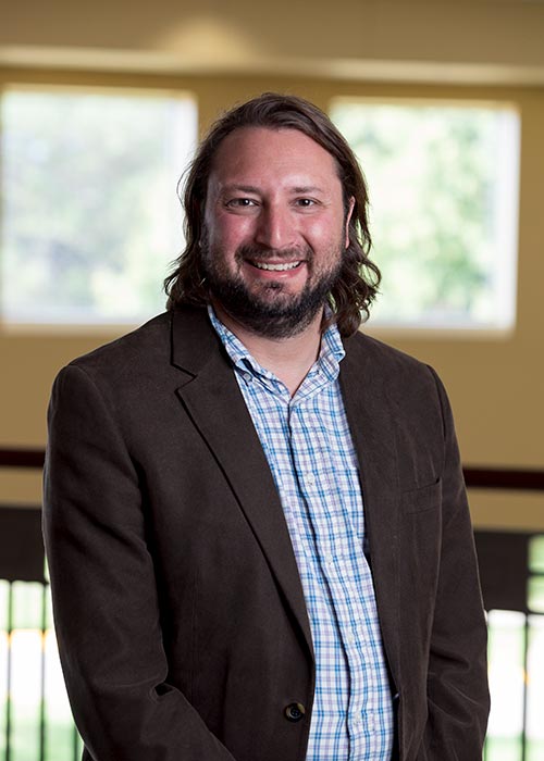 A smiling man with long hair and a beard is wearing a brown jacket over a blue and white checkered shirt. He stands indoors with two blurred windows in the background allowing natural light to enter.