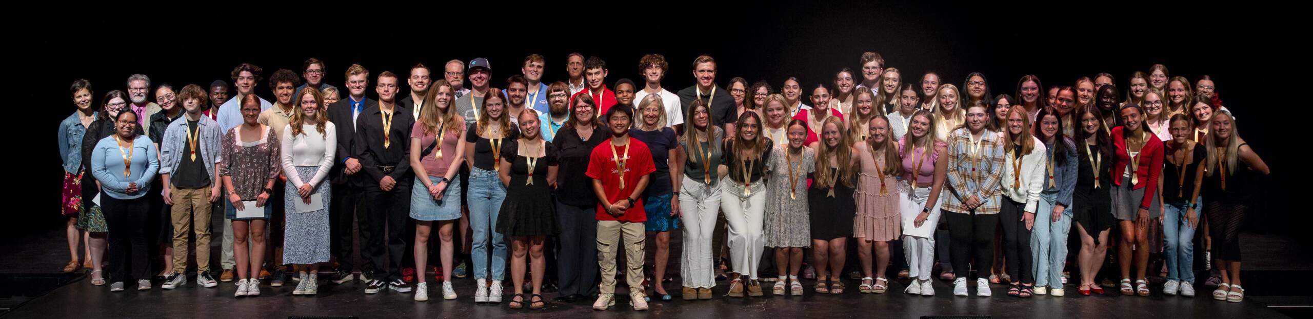 A large group of people, dressed in semi-formal and casual clothing, stand together on a stage against a black background. Many are smiling, and some wear medals around their necks.