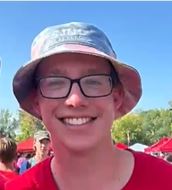 A person wearing a bucket hat and glasses is smiling outdoors. They are dressed in a red shirt, and there are red tents and trees in the background under a clear blue sky.