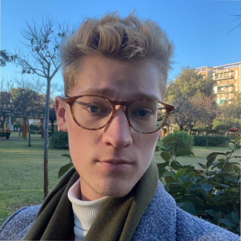 A person with blond hair and glasses is outdoors in a park. They are wearing a coat and scarf. The sun is shining, and there are trees and buildings in the background.