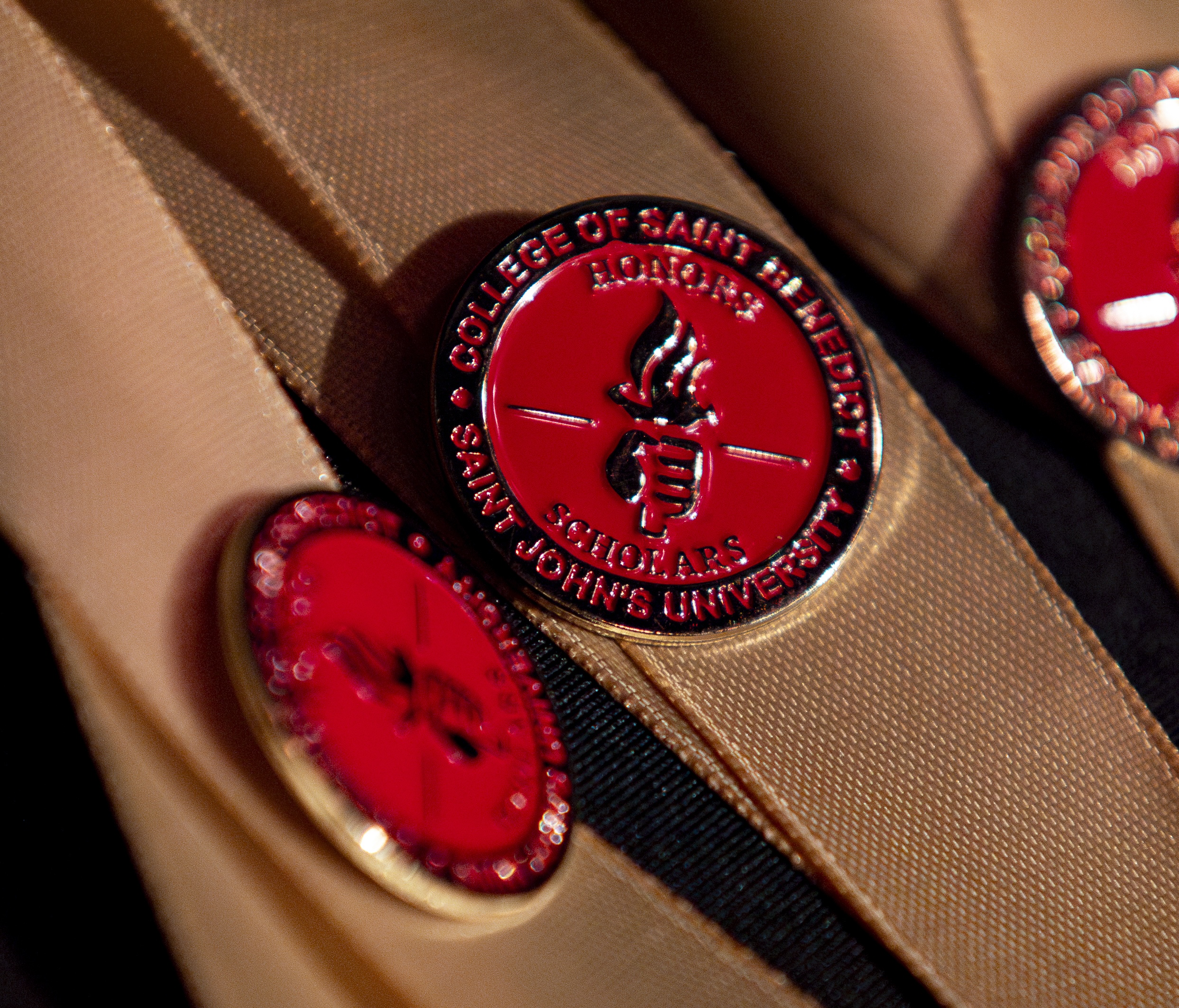 Close-up of a red and gold pin on a tan fabric, featuring the text "College of Saint Benedict" and "Saint John's University Honors Scholars" with a torch emblem in the center. Additional similar pins are partially visible in the background.