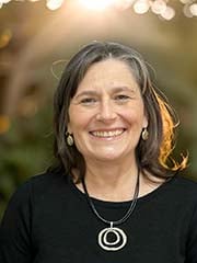 A woman with shoulder-length brown hair, wearing a black top, silver earrings, and a circular pendant necklace, smiles outdoors with sunlight shining behind her.