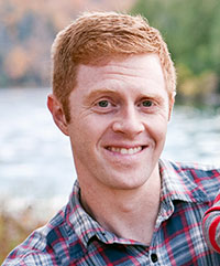 Smiling man with short red hair wearing a plaid shirt, standing outdoors with blurred trees and water in the background.