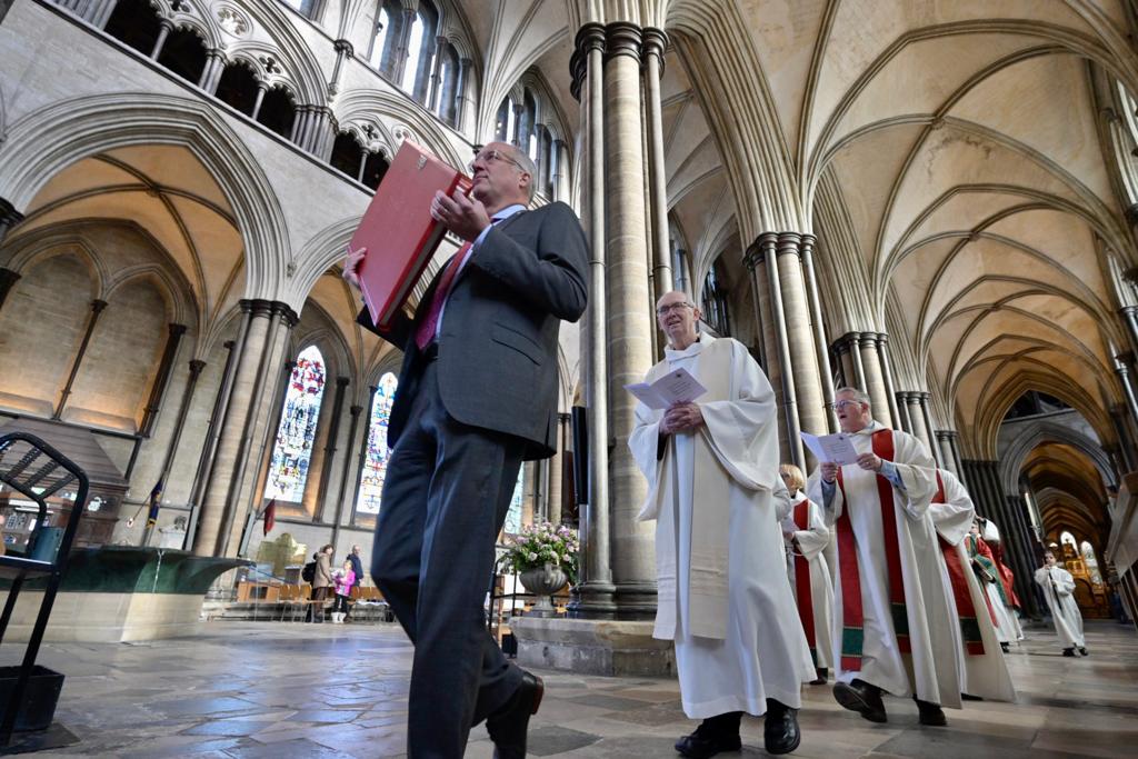 Man carrying Saint John's Bible in a church walking with religious figures walking behind him