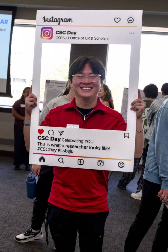 A smiling person in a red shirt holds a large Instagram-style photo frame at an indoor event. The frame reads “CSC Day, Celebrating YOU. This is what a researcher looks like!” with hashtags #CSCDay and #csbsju.