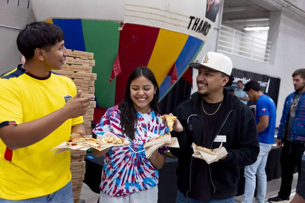 Three young adults stand indoors eating pizza and smiling. One holds multiple pizza boxes, and the others hold paper plates. Colorful backgrounds and people are visible behind them, suggesting a lively social event.