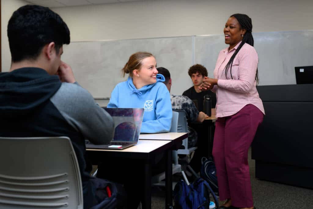 A teacher stands and smiles while speaking to a group of students seated at desks in a classroom. One student in a blue hoodie smiles back, and others are focused on their laptops or listening.