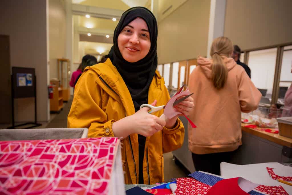 A young woman wearing a yellow jacket and black hijab smiles while cutting red fabric with scissors at a crafting table, surrounded by colorful fabric pieces. Other people work in the background.