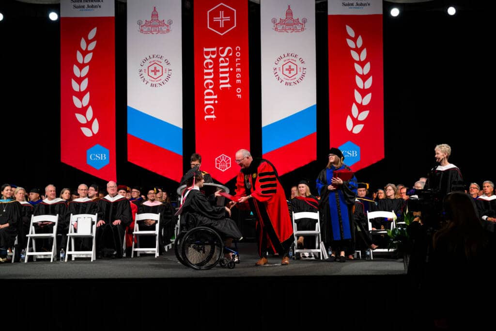 A graduate in a wheelchair receives a diploma on stage during a College of Saint Benedict commencement ceremony, surrounded by faculty in academic regalia and large red banners in the background.