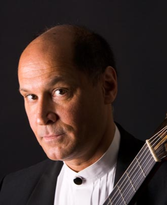 A man with a serious expression is holding a guitar. He has a bald head with some hair on the sides and is wearing a black suit with a white shirt. The background is dark.
