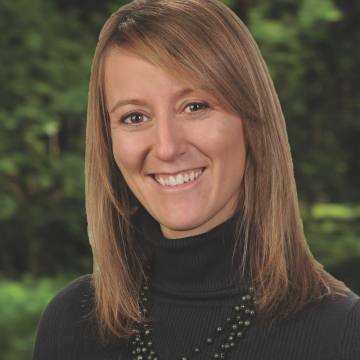 A person with long brown hair is smiling and wearing a black turtleneck sweater and a beaded necklace. The background is a lush green forest, slightly blurred.