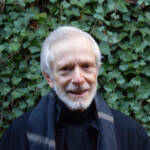 An elderly man with white hair and a beard smiles while standing in front of a wall covered in green ivy. He wears a black coat and a dark scarf.