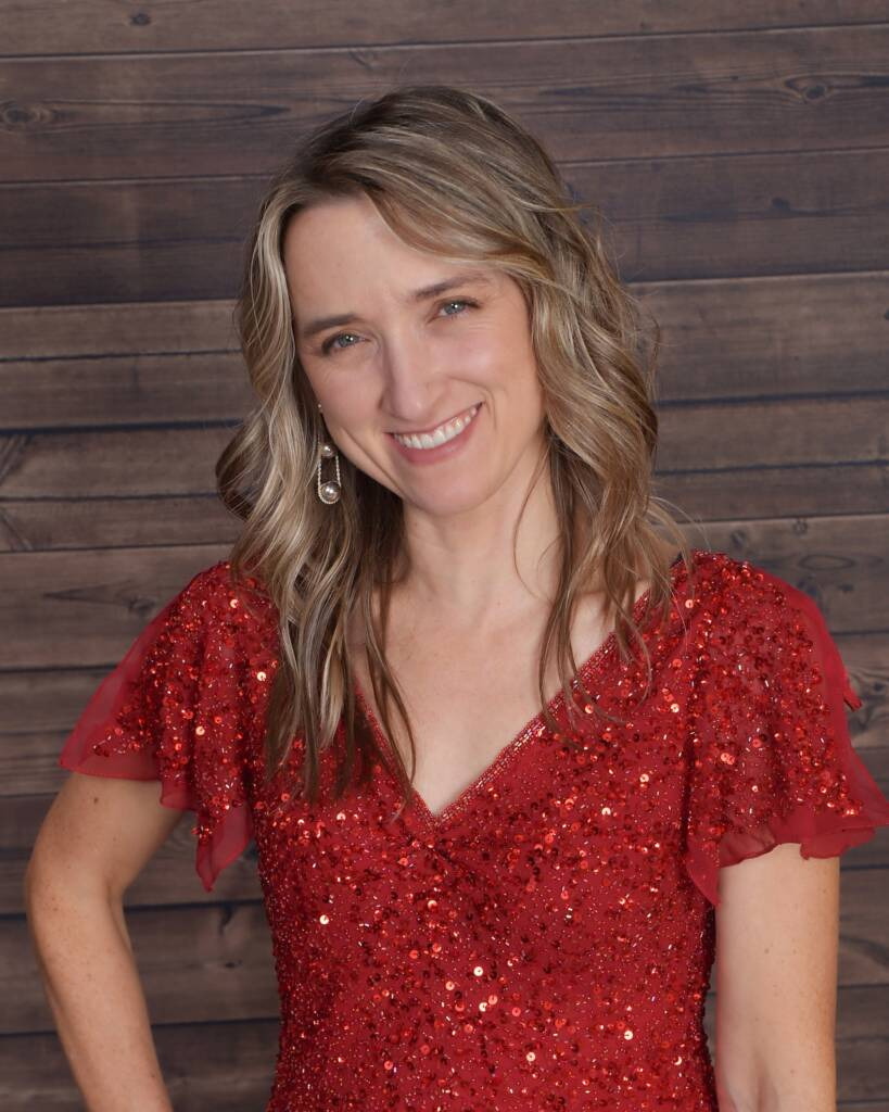 A woman with long, wavy hair and wearing a red sequin dress smiles in front of a wooden wall backdrop.