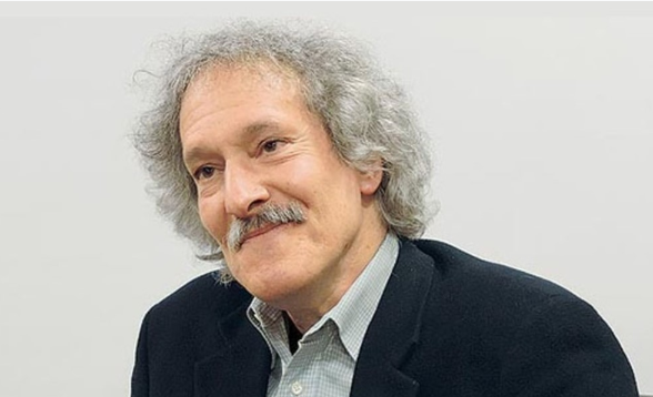 An older person with curly gray hair and a mustache, wearing a dark blazer and light-colored shirt, is smiling against a plain background.