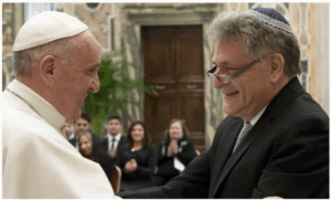 The Pope and a man wearing a kippah smile and shake hands in a formal room, with people applauding in the background.