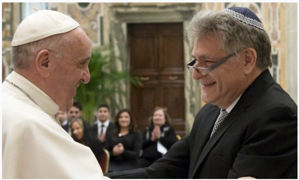 The Pope and a man wearing a kippah smile and shake hands in a formal room, with people applauding in the background.