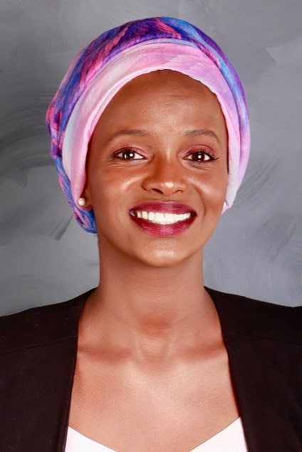 A woman smiles at the camera, wearing a colorful headscarf in pink and blue tones, a black blazer, and a light top. The background is a soft, grayish abstract pattern.