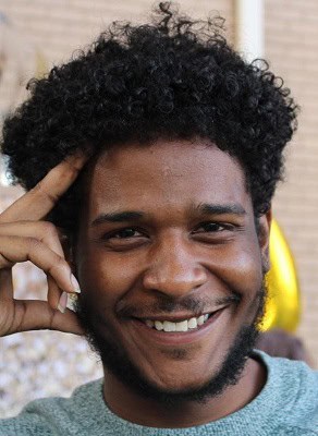 A smiling person with curly hair and a beard rests their fingers on the side of their face. They are wearing a light blue sweater, and the background is softly blurred.