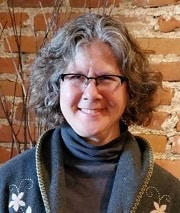 A smiling person with curly gray hair and glasses stands in front of a brick wall, wearing a dark turtleneck and a sweater with embroidered flowers.