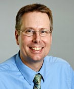 A middle-aged man with short light brown hair, glasses, and a friendly smile, wearing a light blue shirt and a patterned tie, poses against a plain light background.
