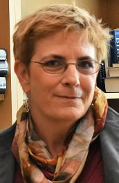 A person with short, light brown hair and glasses is wearing a colorful scarf and a gray jacket, standing in front of shelves filled with books.