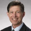 A middle-aged man with short brown hair wearing a dark suit, white shirt, and patterned tie, smiling at the camera against a plain gray background.