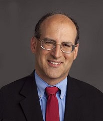 A middle-aged man with glasses, short dark hair, and a receding hairline smiles while wearing a dark suit, light blue shirt, and red tie against a plain dark background.