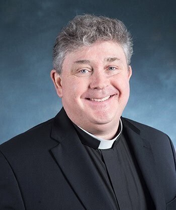 Smiling man with curly gray hair wearing a black clerical suit and white collar, posing against a blue background.
