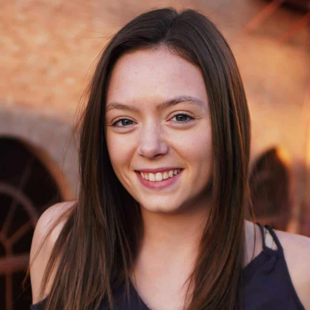 A young woman with long brown hair and a dark sleeveless top smiles at the camera, standing outdoors in front of a blurred brick building and arched windows, with warm sunlight illuminating her face.