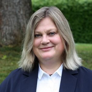 A woman with shoulder-length blonde hair wearing a dark blazer and a white shirt smiles outdoors, with a tree and greenery in the background.