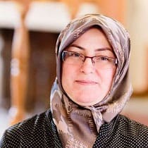 A woman wearing glasses and a patterned headscarf smiles softly at the camera. The background is softly blurred, highlighting her face.