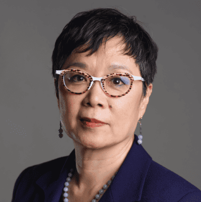 A woman with short black hair, wearing tortoiseshell glasses, a navy blazer, and a beaded necklace, posing in front of a plain gray background.