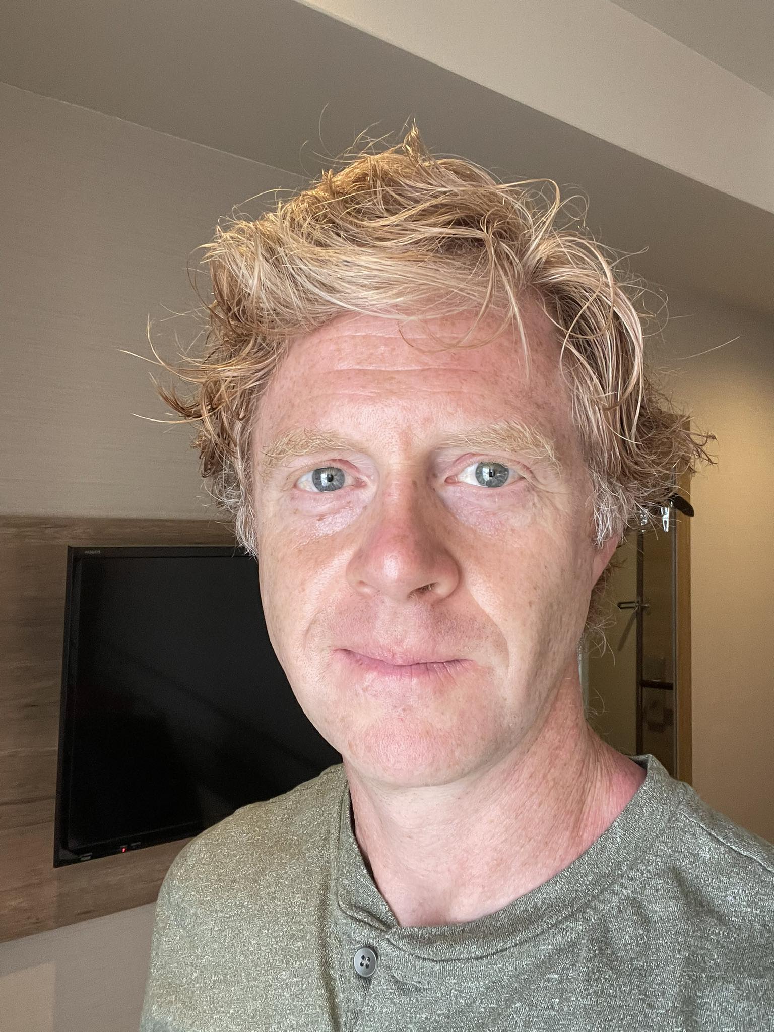 A man with fair skin and wavy blond hair takes a selfie indoors, standing in front of a wall-mounted TV. He is wearing a green shirt and looking directly at the camera with a neutral expression.